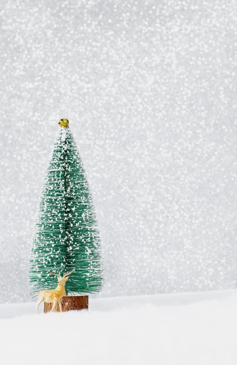 Deer figure in front of a model Xmas tree, with a glittery, snowy background 