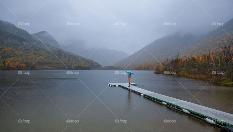 Echo Lake in rain, with clouds and mist lingering with mountains in morning. Young woman standing with a blue umbrella looking into the mountains and lake. Colorful yellow, red and green autumn leaves around. Franconia Notch, NH, US. Nature