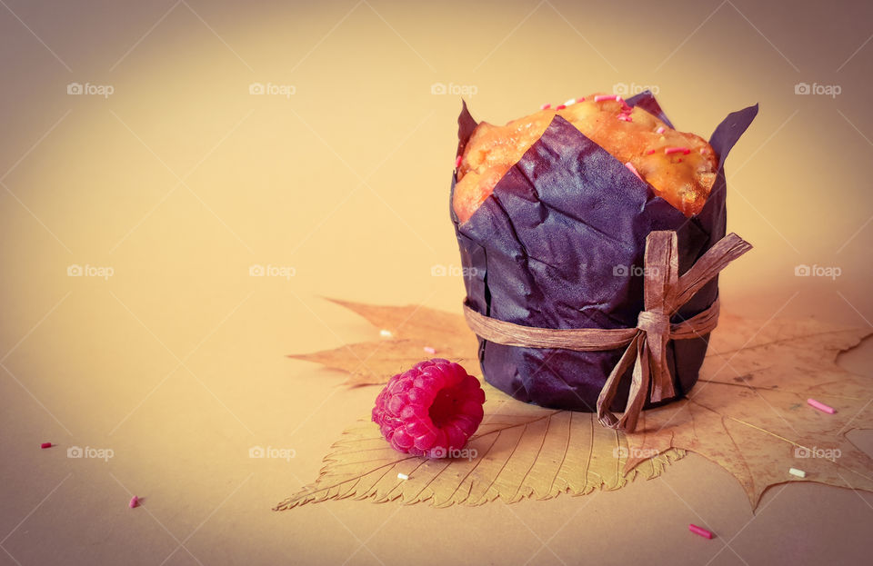 Autumn still life with pastries. Muffin in brown baking paper with raspberries on autumn leaves with pink topping on a plain background. Beginning of autumn