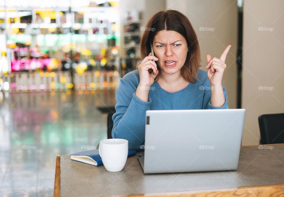 Beautiful stylish young woman plus size body positive screaming over mobile phone using laptop at beauty salon office, small business owner