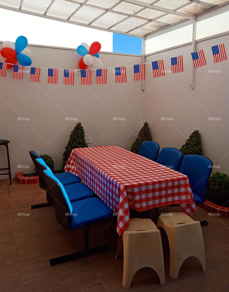 A string o American flags for decoration,  baloons,  checked picnic table,  blue seata, city backyard