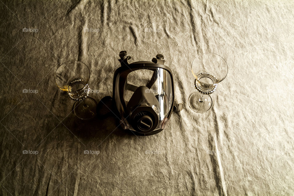 A gas mask rests in between two wine glasses laying on a paint drop against the daytime sun.