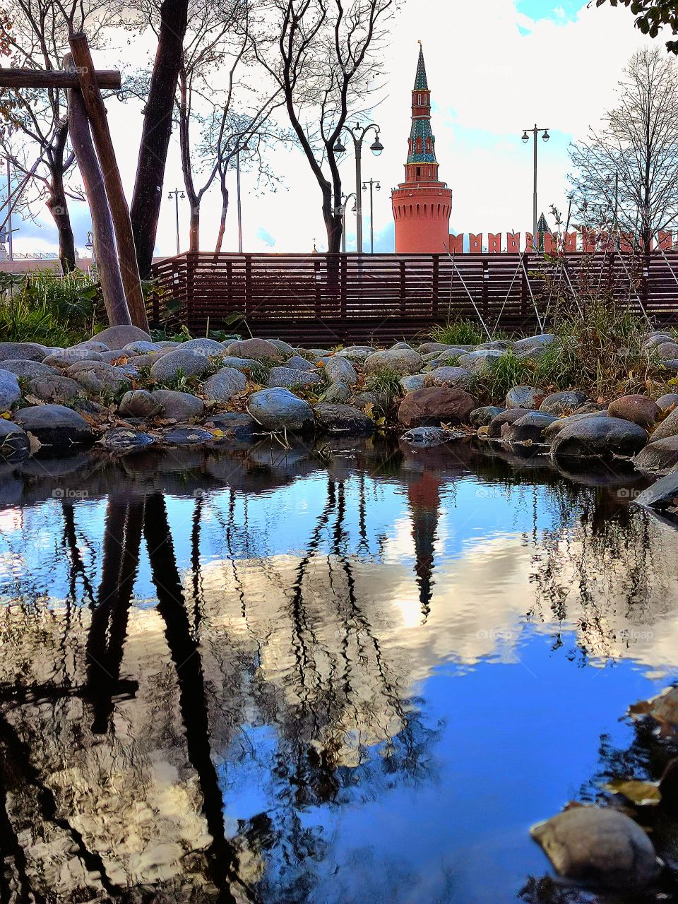 Reflection.  The water of the pond reflects the Beklemishevskaya tower of the Moscow Kremlin, the blue sky with white clouds and trees.  The pond is lined with stones on which fall leaves lie