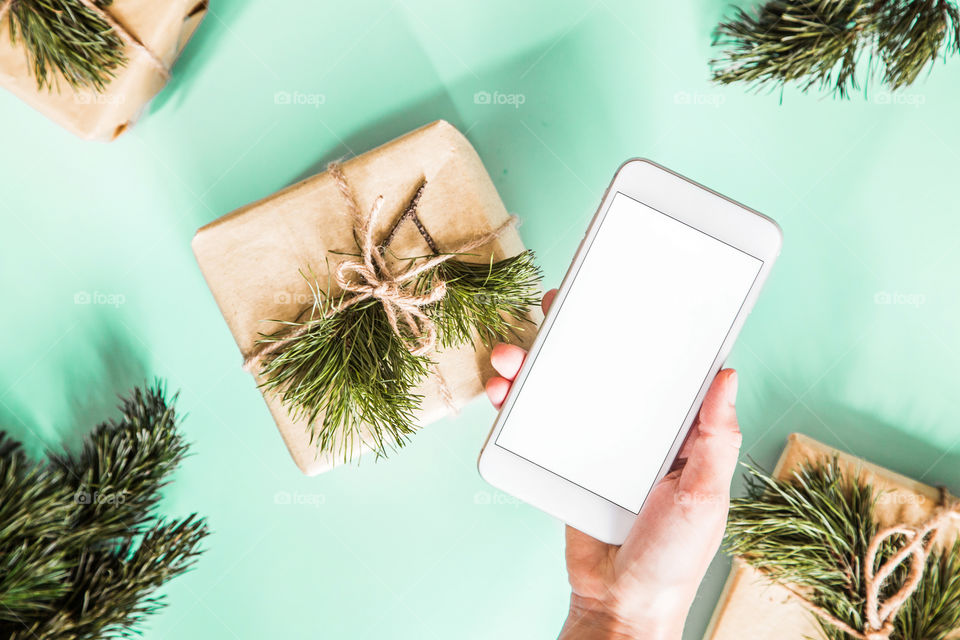 Christmas gift lying on green background. Female hand holding mobile phone. Flatlay items. Flat lay, top view, surprise, happy new year, merry, tree, packing, packed, wrapping, celebrating, holidays, winter, background