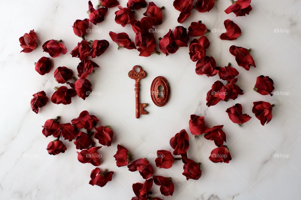 Red, metal, vintage key and keyhole with red, silk rose buds arranged in hear shape on white marble surface
