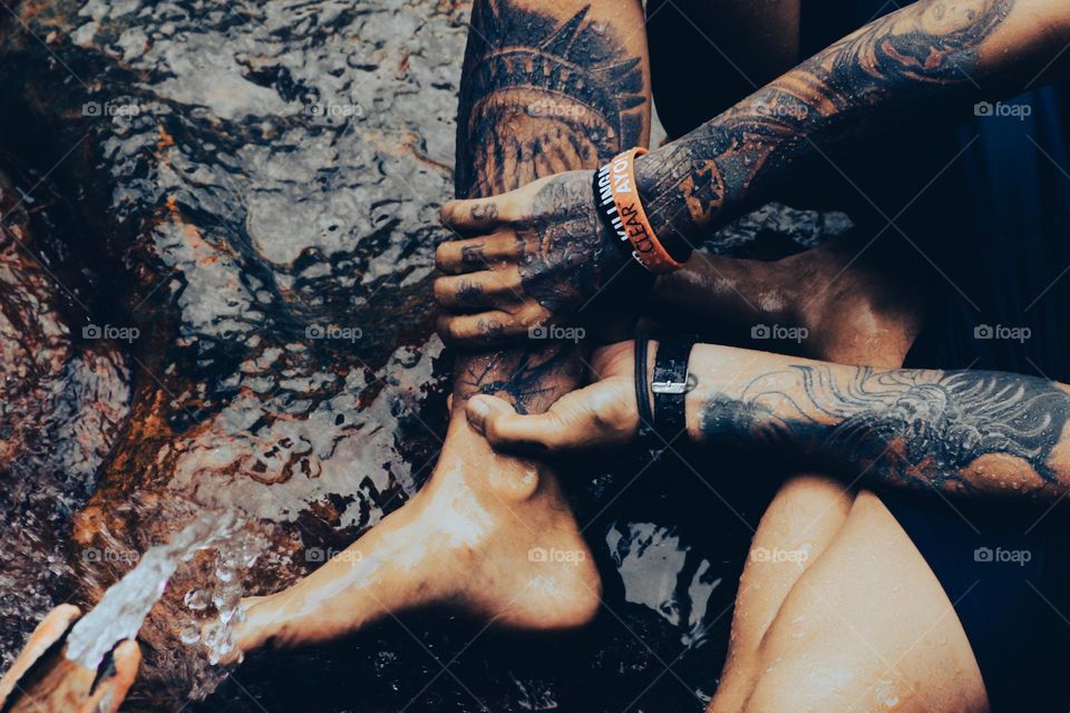 Under a bamboo shower, a tattooed man sits on a mossy rock holding his right leg, both hands and feet covered in tattoos.