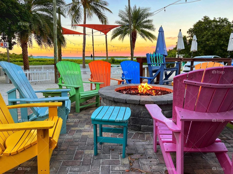 Colorful chairs around a fire pit on a beach at sunset with palm trees