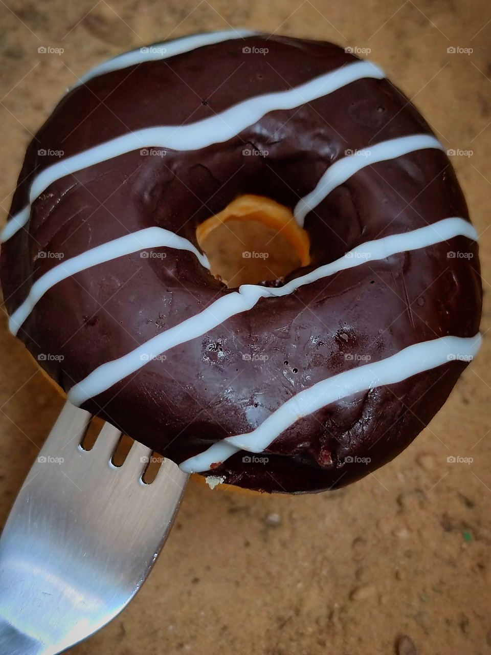 On a fork is a chocolate donut with stripes of white glaze.  Donut on a background of sand