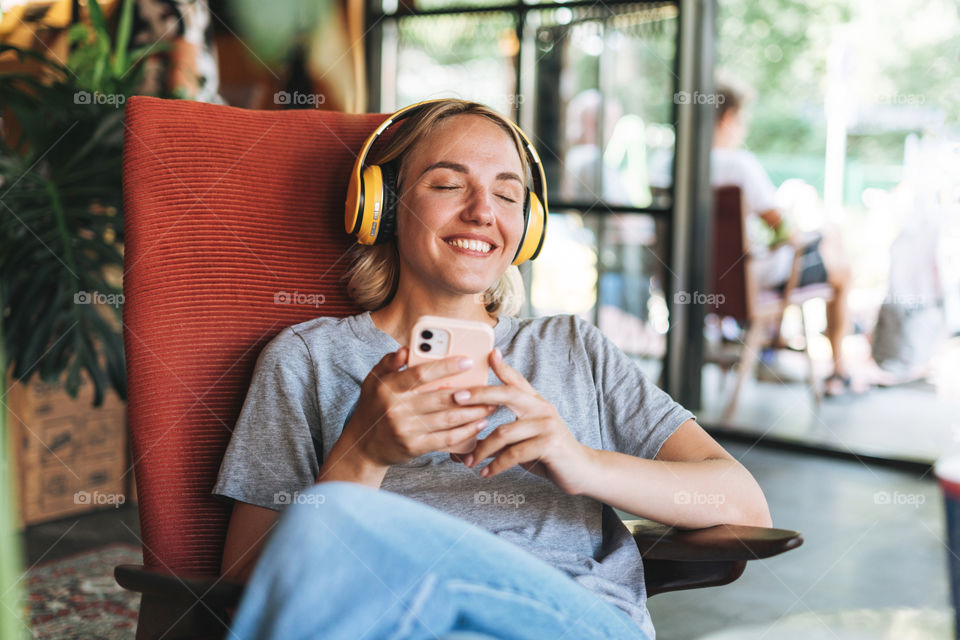 Happy young blonde woman in yellow headphones and grey t-shirt listening music from mobile phone in cafe 