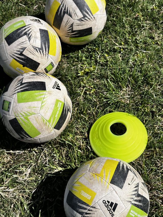 Four Adidas Tsubasa training soccer balls with green and yellow design with fluorescent yellow/green training cone at a practice on grassy soccer field