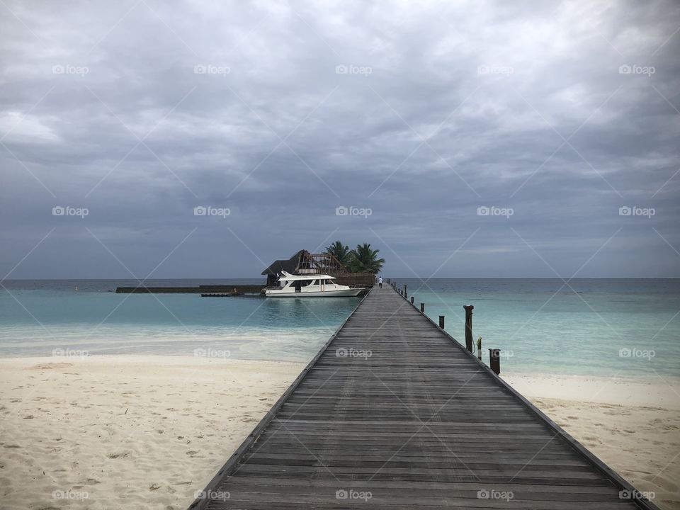A long dock extends to a yacht and several palm trees, under a cloudy gray sky, hovering over blue water and white sand. 