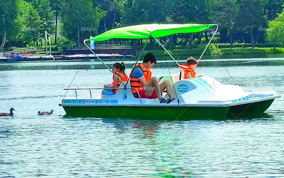 Summer in the city. Pond in the park. A catamaran with a sun-protective green roof floats on the water. Father, son and daughter are sitting in the catamaran. Ducks follow the catamaran