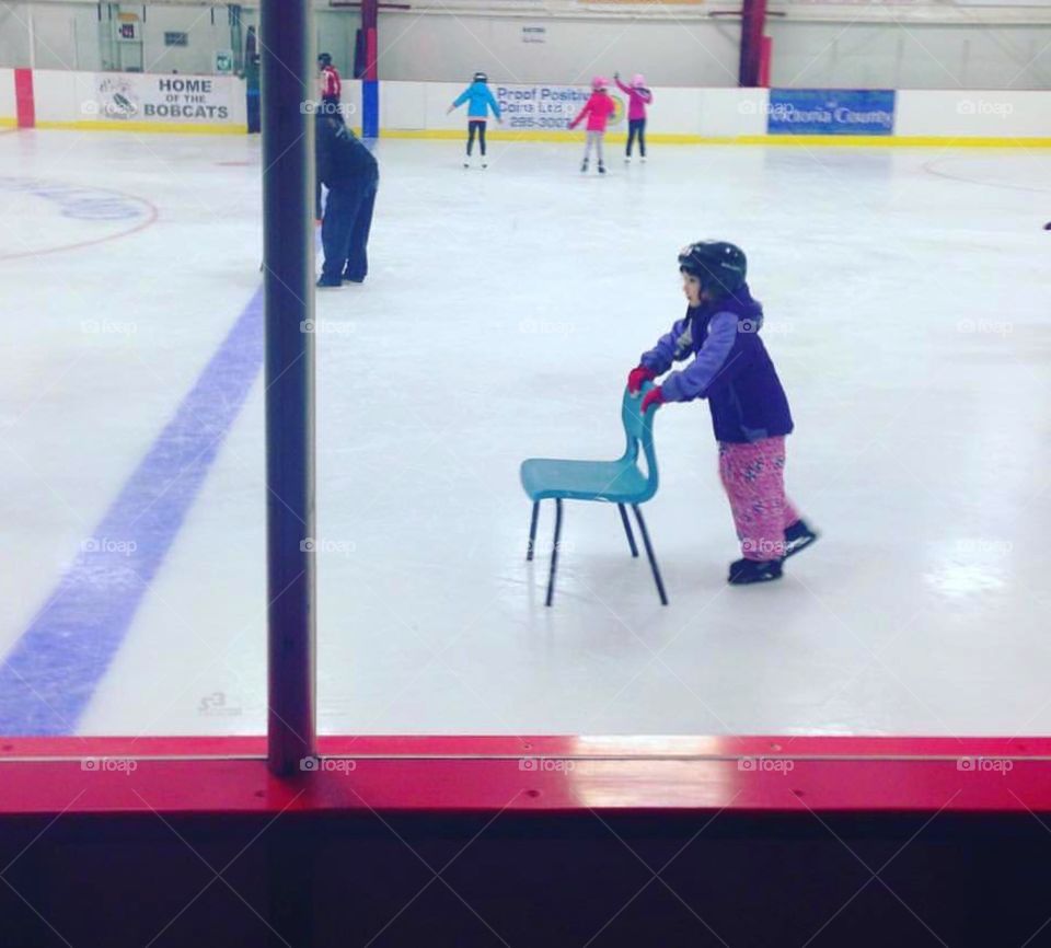 Little girl pushing a chair while learning to skate at an ice rink. Other people skating in background. 