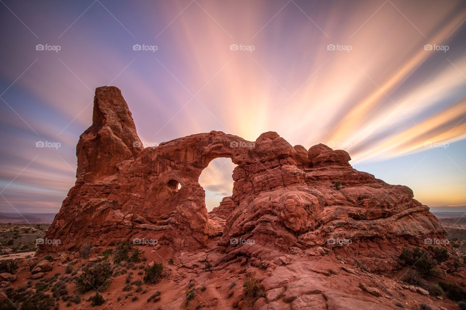 Colorful clouds streaking across the sky above a beautiful sandstone arch, bathed in golden hour light during a sunset.