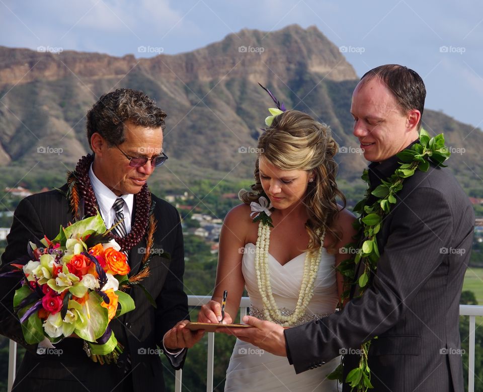 Exterior daylight.  Honolulu, Hawaii, USA.  Foreground:  a bride and groom sign their wedding certificate.  Background:  Diamond Head.