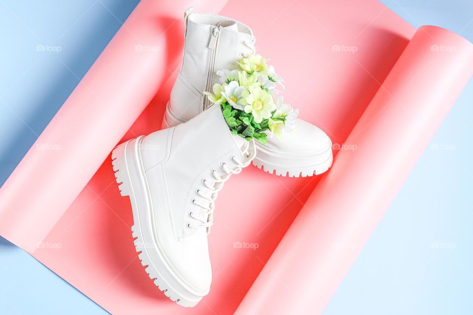 Beautiful white boots on a rough tractor sole with lacing and a faux bouquet of spring flowers on a rose blue background,flats Lay. The concept of fashionable women's shoes.