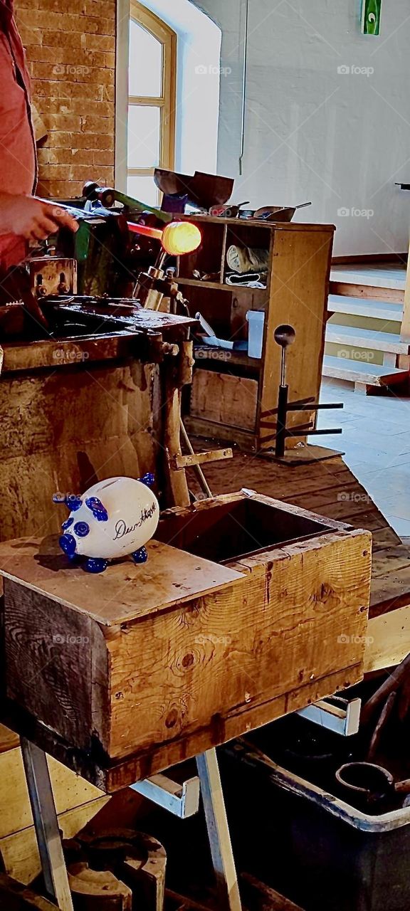 In “Freyung”, Bavaria a glowing hot piece of raw glass is being manipulated on one end of a long thin pipe that will later carry air into the solid mass making it hollow and shaping it. 2023. Hypnotic Productions