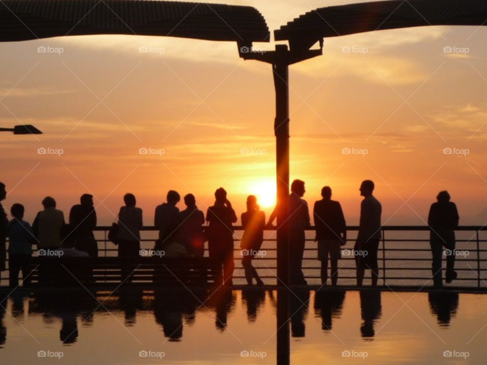 people from behind contemplating the sunset from a railing reflected in the water of a fountain