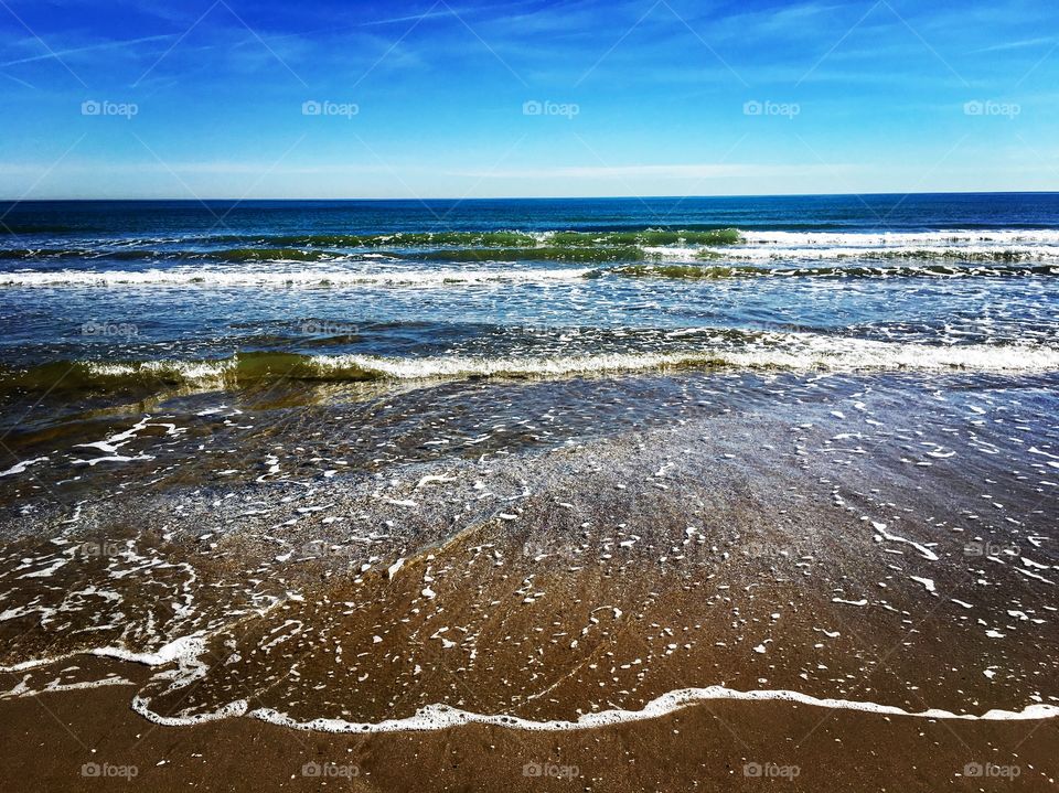 Small waves crash into a brown sand beach on a warm spring day. 