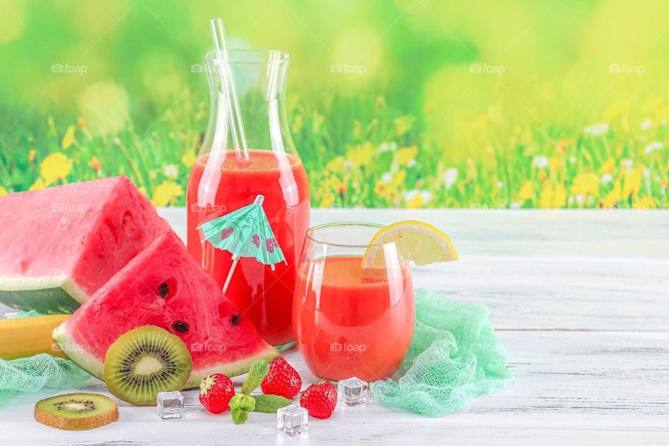 Multivitamin red drink in decanter with glass, fresh slices of watermelon, kiwi, lemon, strawberries, rosemary twigs, ice cubes, glass straws and skewer paper umbrella on white wooden shabby boards against a blurred background of a flowering field, c