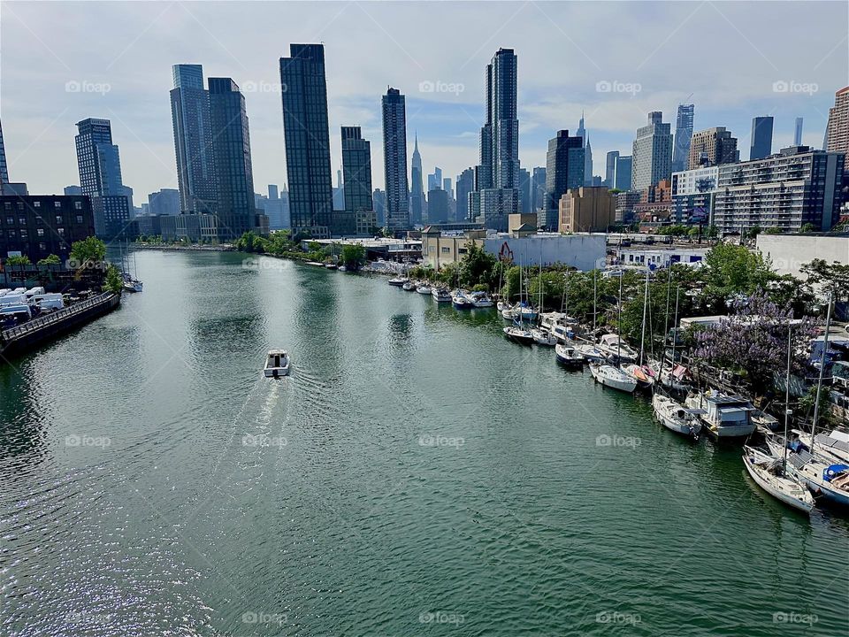 This is “Newtown Creek” with its boats on the water as well as on the shore seen from the “Pulaski Bridge” that connects LIC to “Greenpoint”, Bklyn. Across the “East River” we see “Manhattan” incl. the “Empire State Bldg”. 2024. Hypnotic Productions