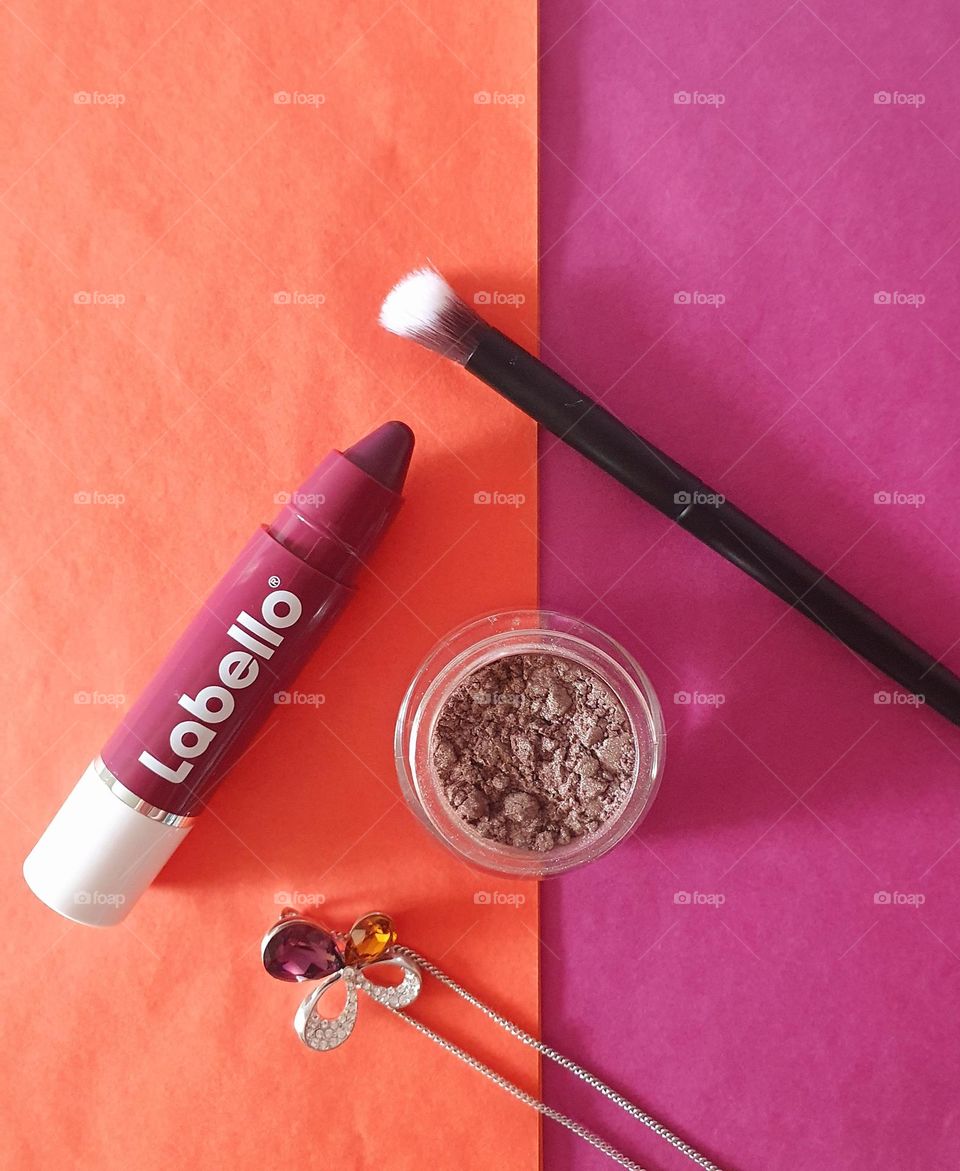Labello black cherry crayon flatlay on an orange and purple paper surface with purple eye shadow, eyeshadow brush and a silver pendant with purple, orange and white crystals