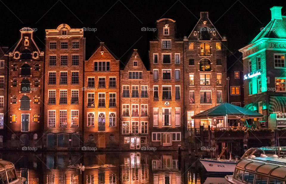 Traditional canal houses in Amsterdam, The Netherlands at night. Colorful night view of waterfront typical Dutch houses facing the Amstel river. Reflections on the water.