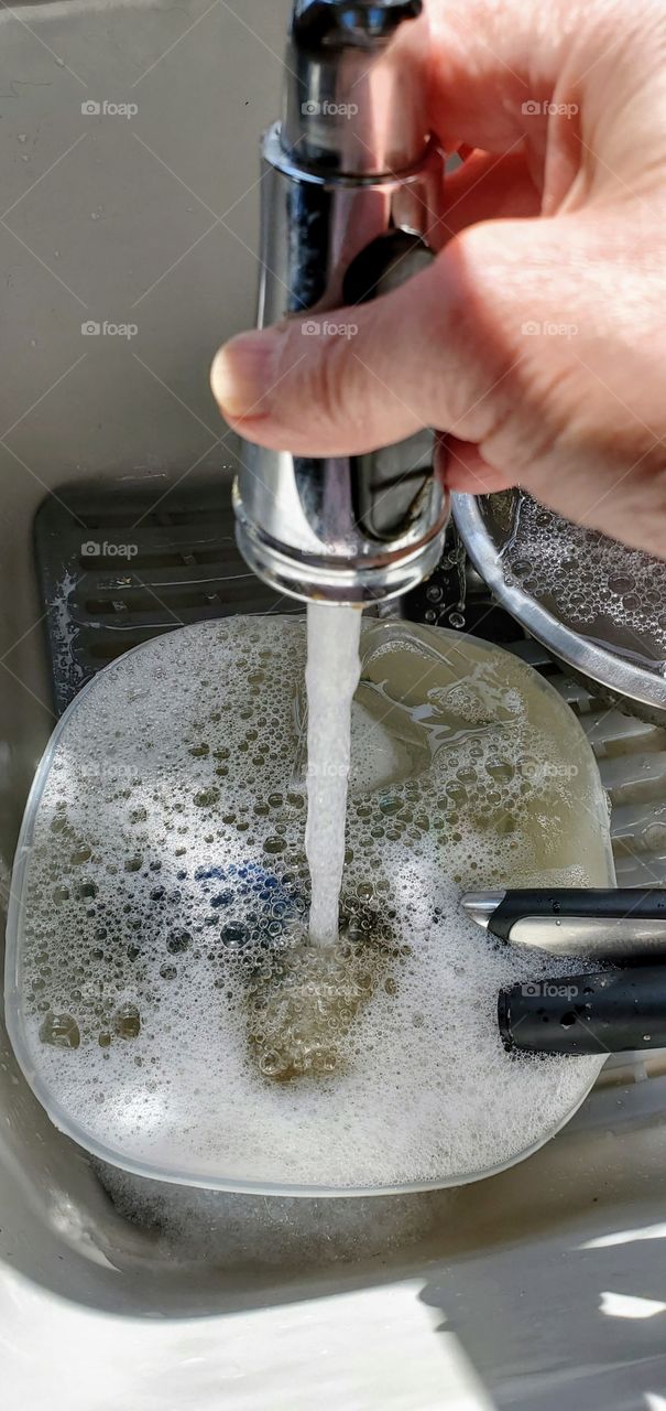 Kitchen sink faucet, running water into bowl creating detergent bubbles. Time to wash the dishes.