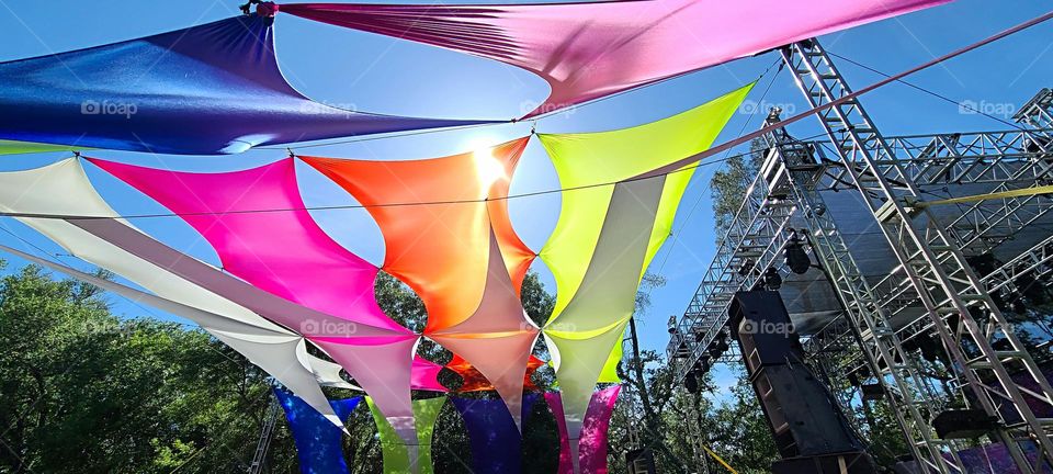 colorful pink, yellow and orange  shade sails with the sunshine and blue skies behind them at a concert