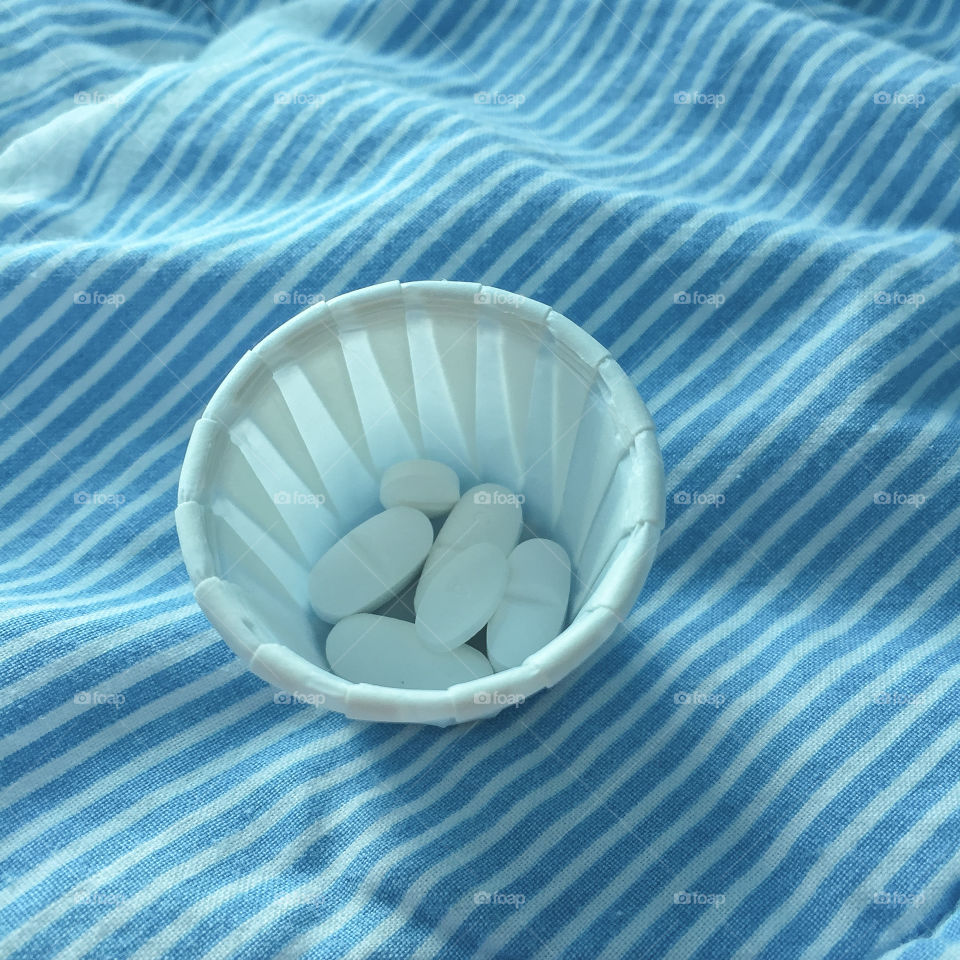 Small paper mug containing a dose of medicine and standing on a striped quilt on a hospital bed.