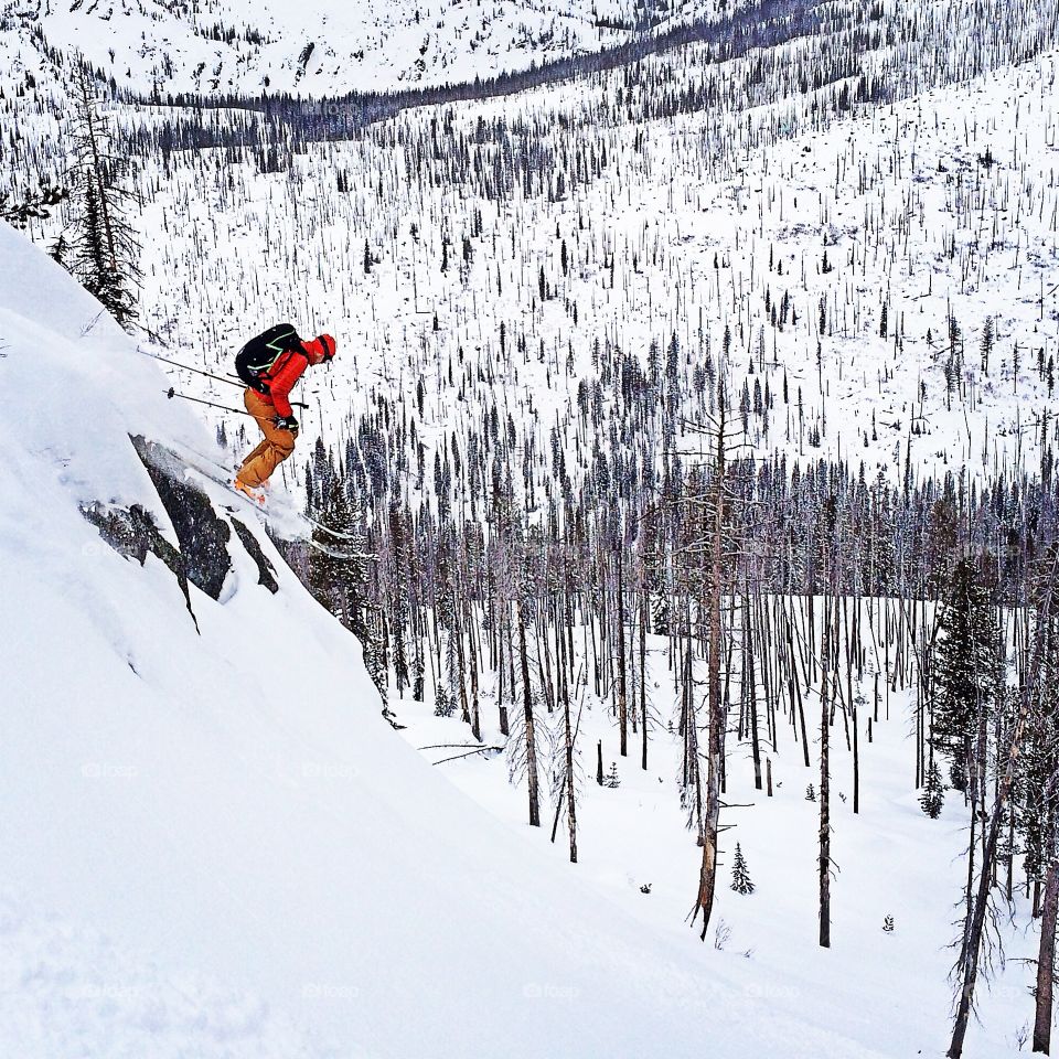 Backcountry skier launching off of rock in Idaho mountains. Colorful kit pops against white landscape. 