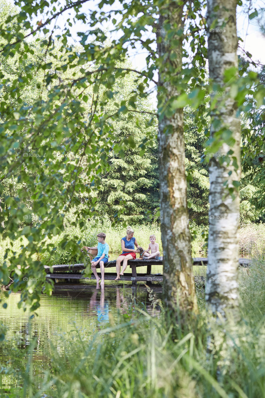 Family spending time together sitting on a bridge over a lake, among the trees, close to nature, during summer vacations. Candid people, real moments, authentic situations