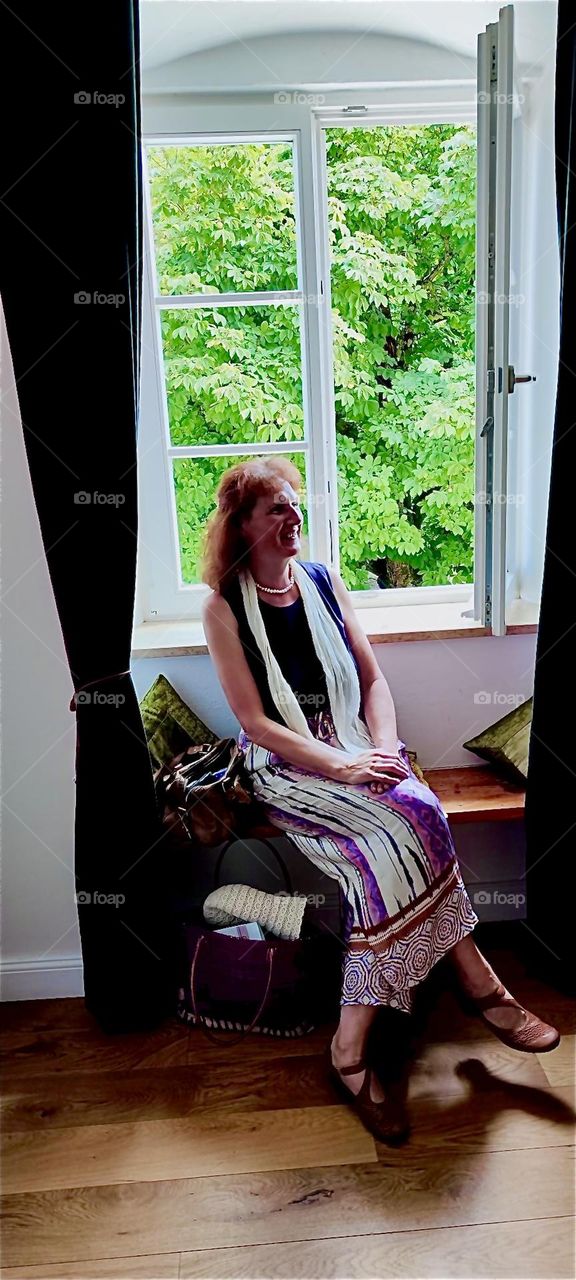 Our lovely lady friend sits inside by the open window of „Schloß Blumenthal“, „Castle Blumenthal“ in „Aichach“, „Bavaria“, Germany waiting to be seated at the gourmet restaurant for hearty Bavarian fare. 2024. Hypnotic Productions