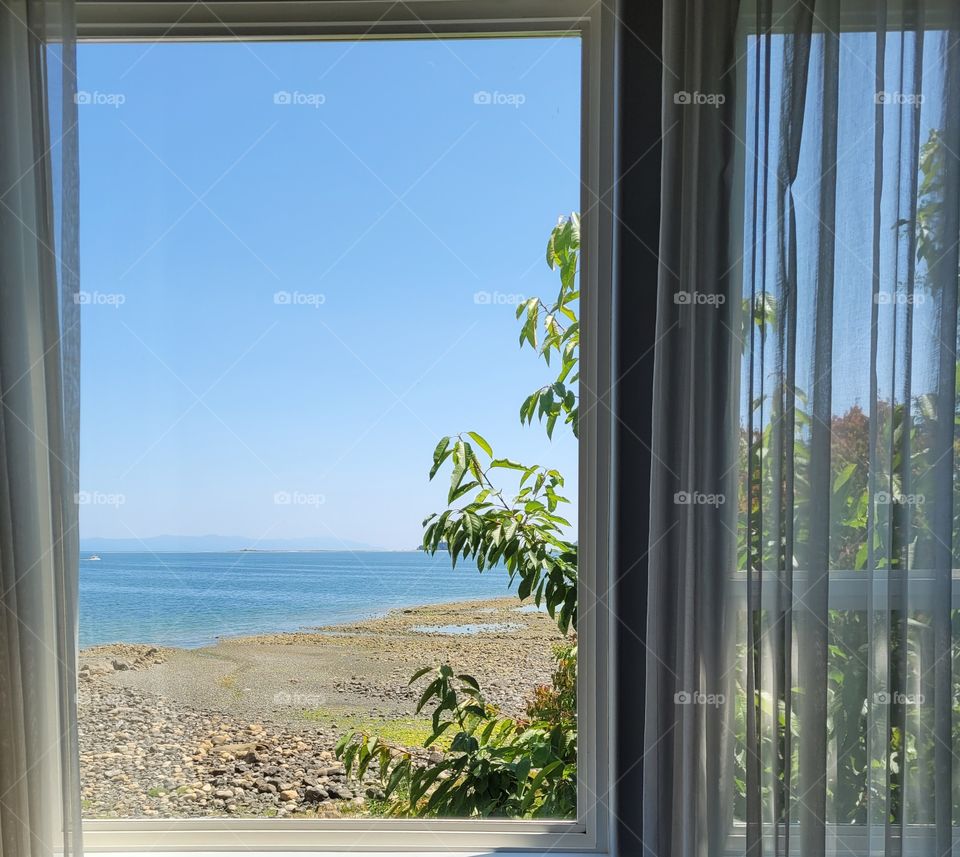 Looking out clear glass window to sunny beach view with blue sky and ocean, with sand and rocks