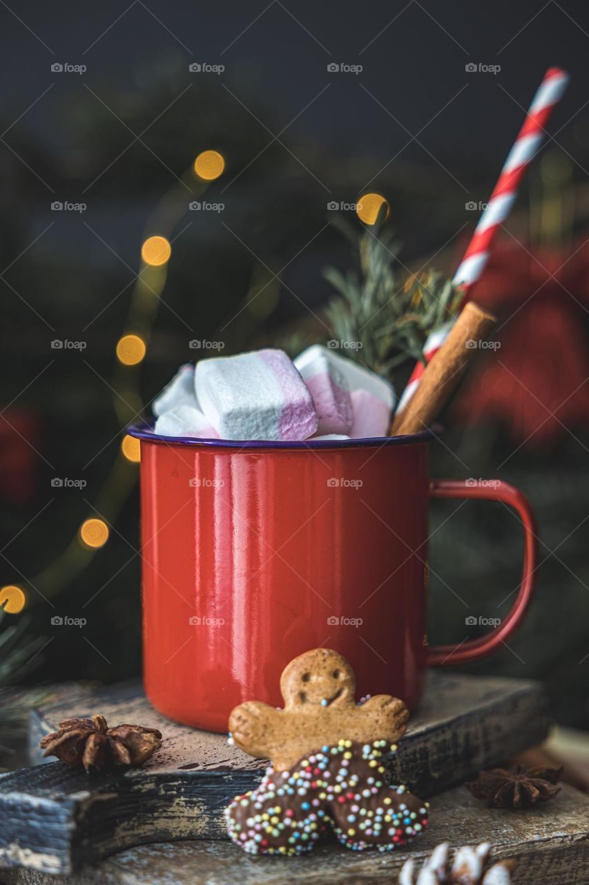 One large metal hot drink mug with cinnamon, marshmallow,straw, anise and gingerbread man cookies on cutting boards with a blurry background of a Christmas tree and burning garland,side view. Holiday Drinks Concept.