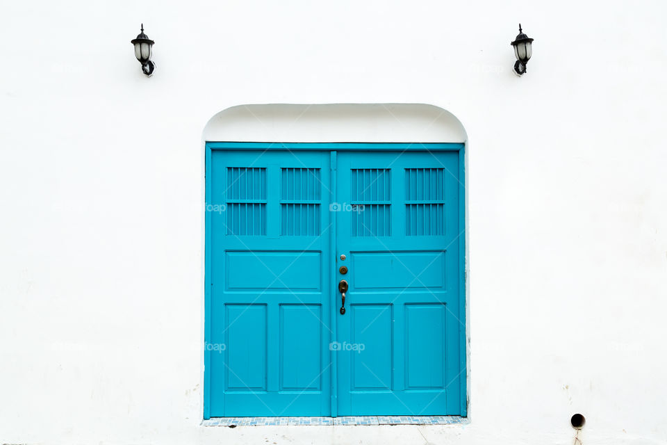 Newly painted light blue wooden double doors on clean white wall. Strong color contrast. Two black metal street lamps