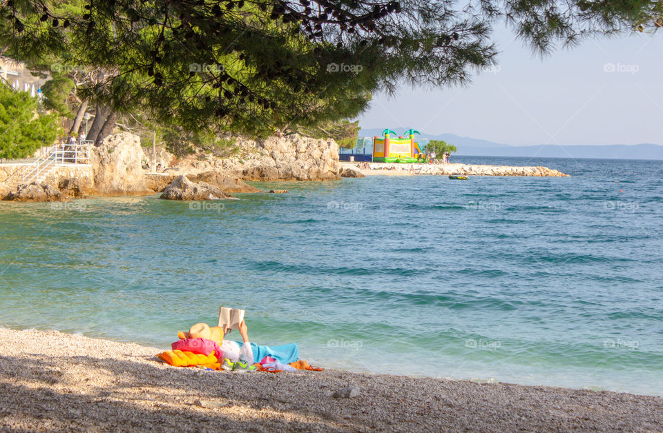 Summer vacation alone, relax. The girl lies on a deserted pebble beach at the edge of a calm sea under the branches of a pine