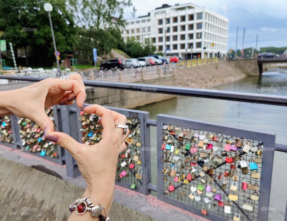 Human hands making heart shape with the bridge with plenty of locks as symbol of love, canal and modern building on the background 