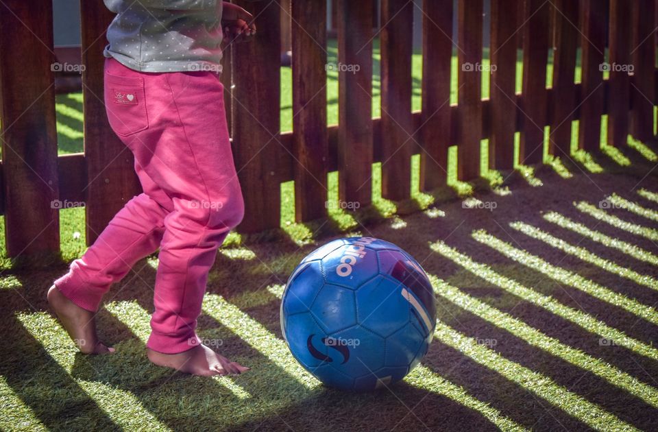 Little girl with pink pants on the playground, kicking a blue soccer ball on green Astro turfed glass