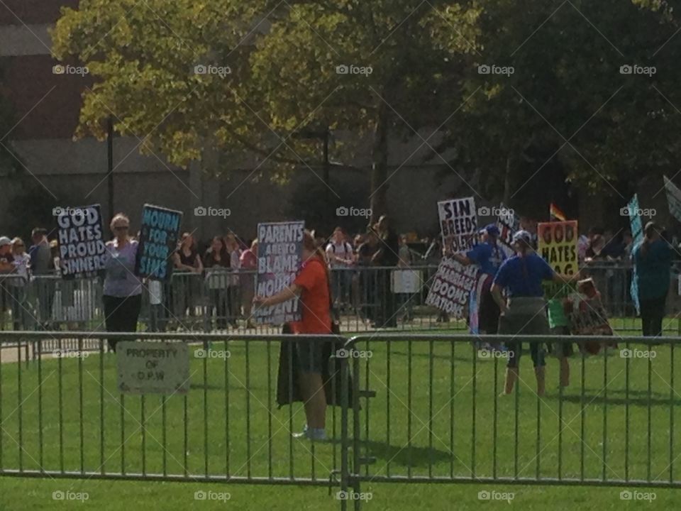 Westboro Baptist church members holding signs to protest an LGB club formed by students of IUPUI.  Club supporters and spectators stand in the background behind the fence.