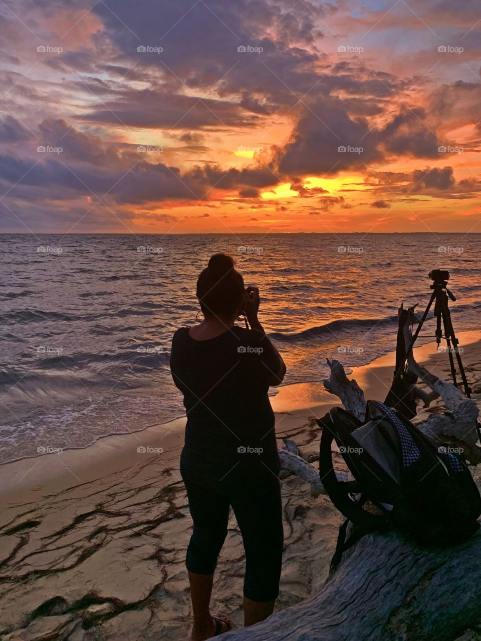 A young lady taking photos of a colorful twilight sunset. The dramatic hurried rush of orange collapse as the sun sank below the gorgeous, cloudy skyline. Waves crash the seashore