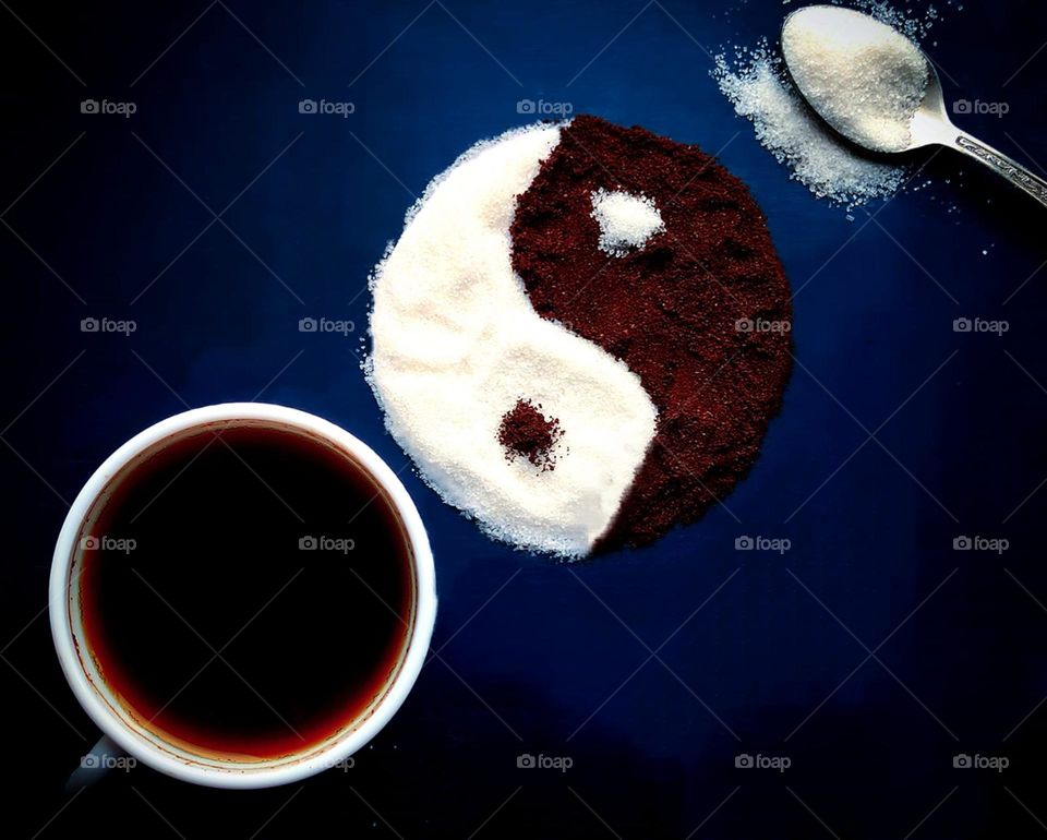 Coffee. The background is dark blue. White cup with black coffee. Yin-yang symbol: white sugar and brown ground coffee. Metal spoon with white sugar