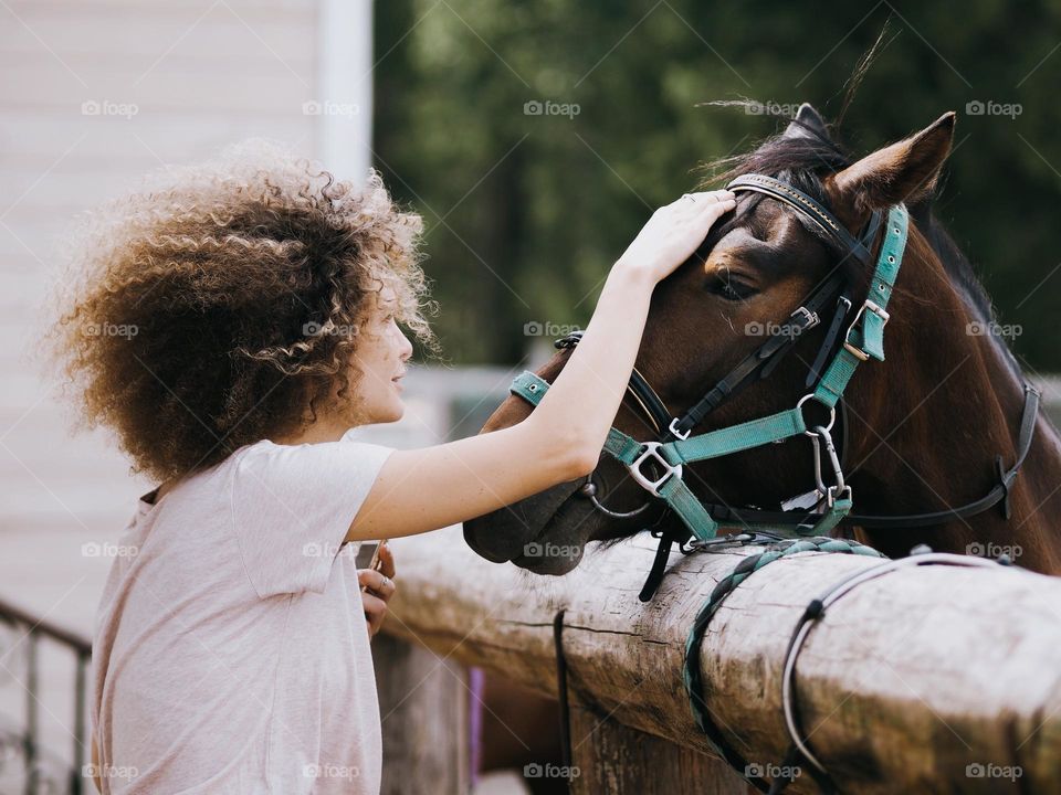 Young beautiful happy girl with curly hair stroking a horse on a farm on a warm sunny day