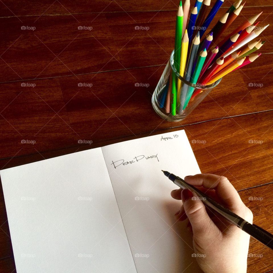 A woman's hand writing in her journal with a glass of colored pencils in frame