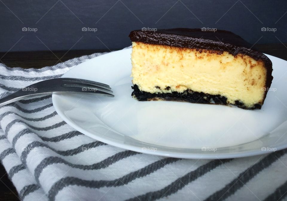 A slice of cheesecake with a chocolate crust, topped with chocolate ganache frosting