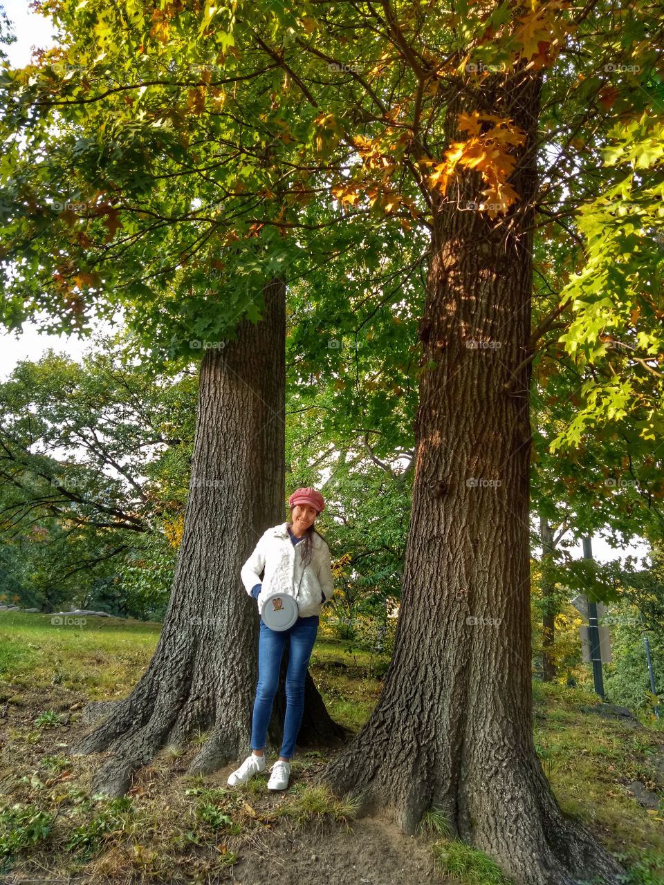 A woman wearing a red hat and white sweater, putting her hands on the pocket. Standing with two tree trunks background. 