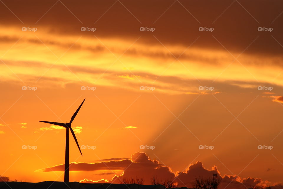 Backlit wind turbine. Orange sky with clouds projecting shadow beams.