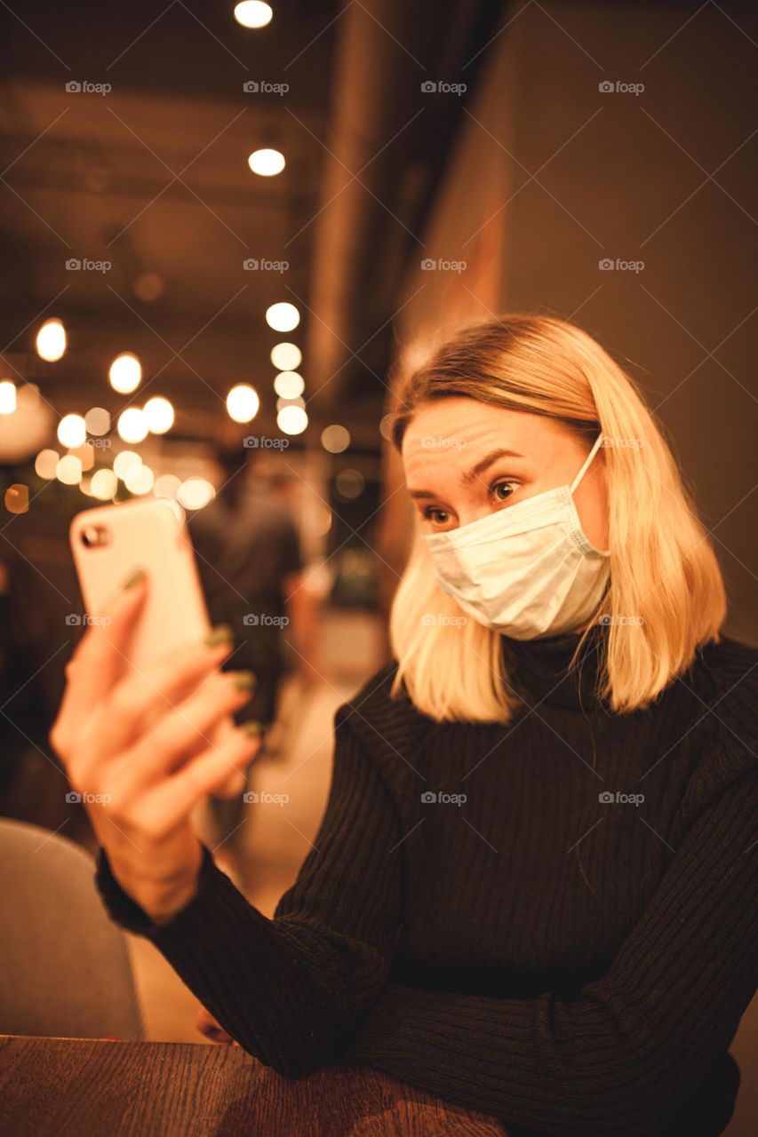 Young woman sits in a cafe, quarantine time. Happy blonde is resting in a cafe, wearing a mask and talking on the phone. Portrait photo of a woman in a black sweater