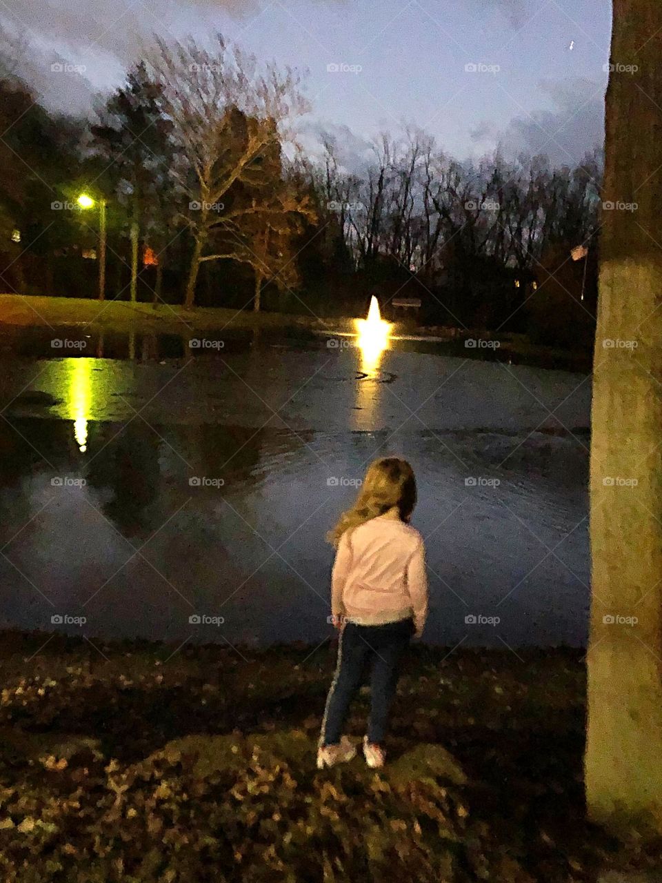 My youngest daughter standing in front of Brookside pond which is lit by the light of the fountain surrounded by trees and streetlights 