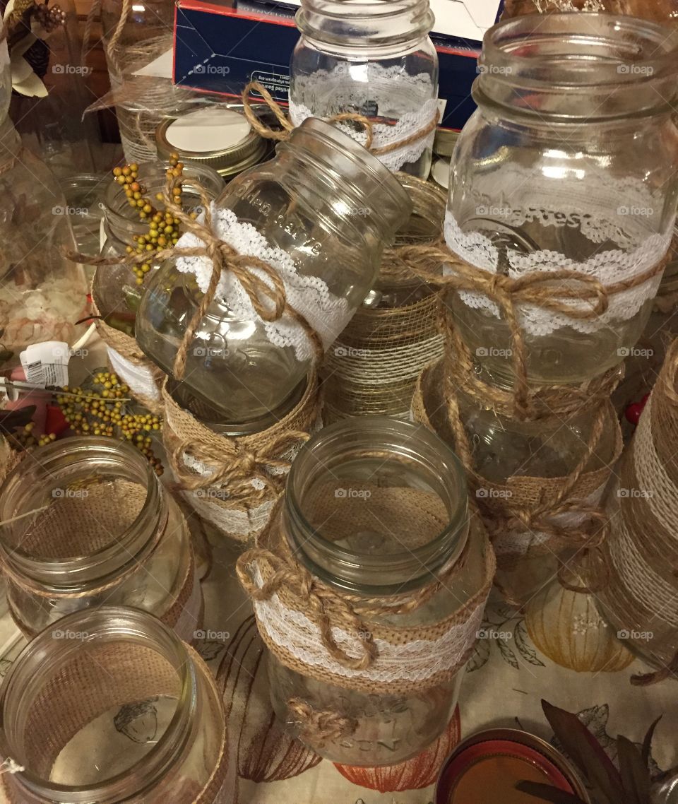 Glass mason jars decorated with burlap , lace, and twine, piled on a table.